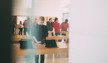 Young female customer standing near table with electronic products while examining the goods, store employees and visitors stand in blur on background. Millennial hipster girl checking device for buy