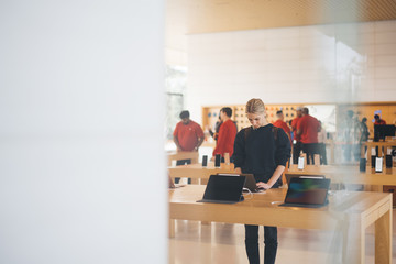 Young female customer standing near table with electronic products while examining the goods, store employees and visitors stand in blur on background. Millennial hipster girl checking device for buy