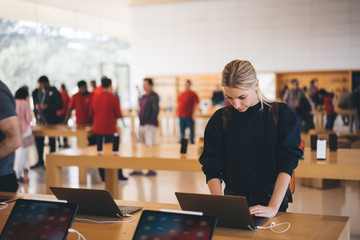 Young female customer standing near table with electronic products while examining the goods, store employees and visitors stand in blur on background. Millennial hipster girl checking device for buy