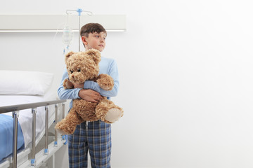 Child in hospital room standing next to bed hugging a teddy bear wearing a pajama isolated on white wall with copy space