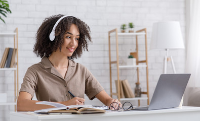 Workplace at home for study and work. African american girl listens online lesson, writes in notebook and looks at laptop