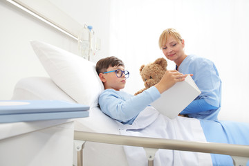 Child reading a book and lying in bed in hospital room with a nurse holding a teddy bear sitting...