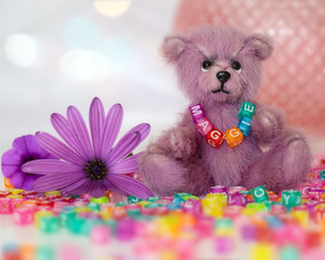 Little pink teddy bear sitting among letter beads and wearing a necklace with the name Maggie