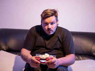 Hombre joven jugando videojuegos mientras come palomitas en el salón de su casa sobre un sofá...