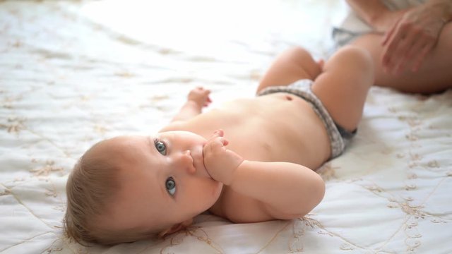 Sweet little baby wearing pants lying on white bed at home and looking at camera. 