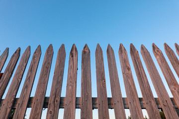 A look from below at an unpainted wooden fence made of pointed slats. The wooden elements are covered with cracks and burrs. The blue sky is visible in the background.