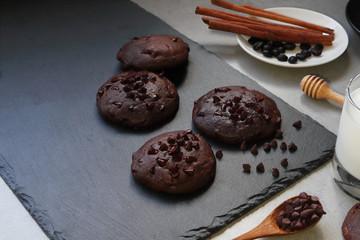 chocolate chip cookies are put on the black stone tray with a glass of milk and decorate with cinnamon sticks and chocolate chips on the grey table in the morning
