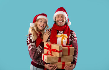Portrait of adorable young couple in Christmas outfits holding heap of gift boxes on blue background