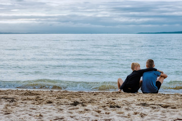 Two young brothers sitting on a beach.  The older brother is looking at the sea and an island on the other side and the younger brother is holding his arm over his older brother.