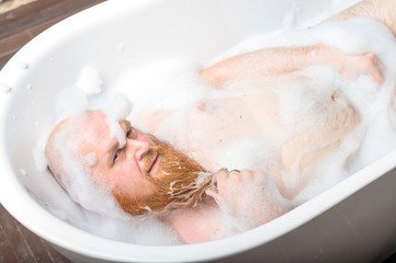 Close-up portrait of funny red-bearded bald man taking a bath with foam. Cheerful naughty guy at spa treatments. A parody of glamorous girls.