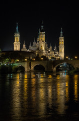 La basílica del pilar de Zaragoza iluminada de noche con el reflejo del puente en el río.