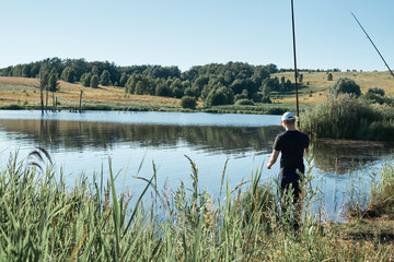 Obraz na płótnie Canvas Summer fishing for fishing rod. A fisherman stands against a background of a beautiful, calm lake with reflection of trees. A beautiful scene of outdoor activities