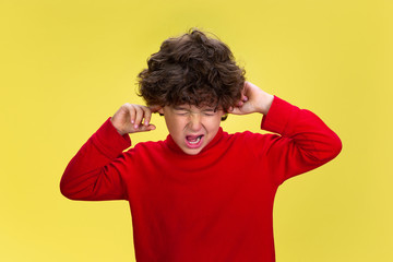 Fototapeta na wymiar Closing ears. Portrait of pretty young curly boy in red wear on yellow studio background. Childhood, expression, education, fun concept. Preschooler with bright facial expression and sincere emotions.