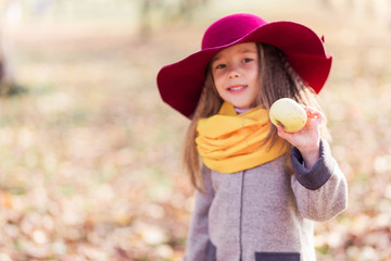 Beautiful little girl in a gray coat, yellow scarf, burgundy hat, with a green apple in her hands posing for the camera in an autumn park