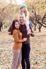 Fototapeta premium Mom, dad and their little daughter are posing in the autumn park. The girl sits on the shoulders of dad