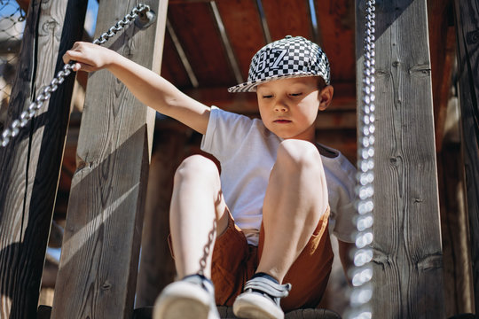 cute caucasian boy in checkered  baseball cap and white tee shirt climbing down the ladder at the wooden playground. He is holding on the chains. Image with selective focus