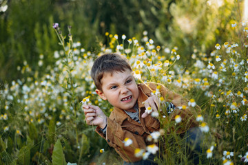 cute caucasian boy in striped henley and brown jean jacket smiling sitting on the glade of wild...