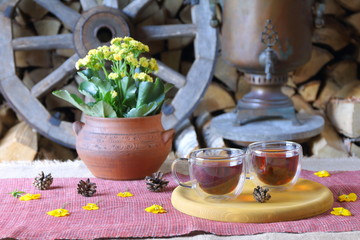 A couple glass mugs of black tea on the background of wooden wheel, samovar and stack of firewood.