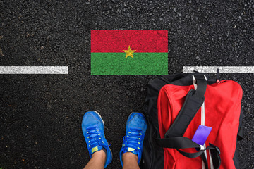 a man with a shoes and travel bag is standing on asphalt next to flag of Burkina Faso and border 