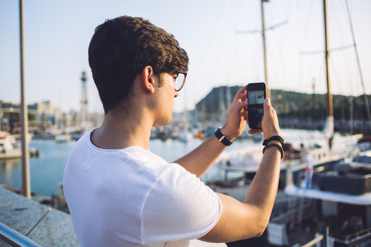 Rear view of male millennial influencer making picture on smartphone camera during trip in harbor, hipster guy taking photo of yachts and boats using mobile phone explore destination in journey