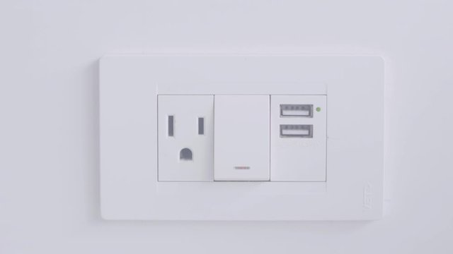 A light switch, a three legged cable plug in and two small usb ports are part of a device placed in the wall of a white room.