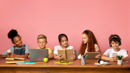 School time. Multiethnic kids with different gadgets and books sitting at table against pink...