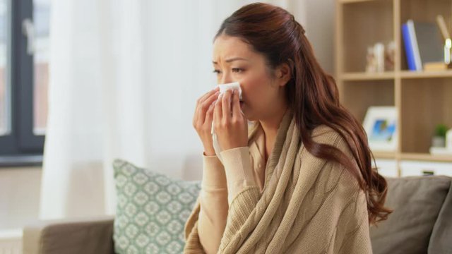 healthcare, cold, hygiene and people concept - sick asian woman in blanket taking paper tissue from box and blowing her nose at home