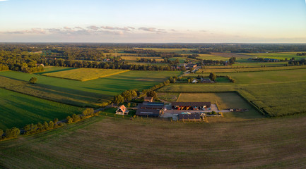 Fototapeta na wymiar Aerial view of a Dutch farm at sunset with warm colours in the low clouds under a blue sky. Agriculture food industry farmland.
