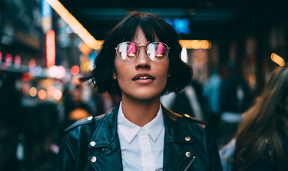 Kaukasische Frau in optischer Brille mit Neonreflexion von Lichtern, die während der Reiseferien in der Urbanität stehen, um New York zu besuchen, attraktives Hipster-Mädchen in trendiger Kleidung beim Ausgehen © BullRun