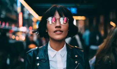 Kaukasische Frau in optischer Brille mit Neonreflexion von Lichtern, die während der Reiseferien in der Urbanität stehen, um New York zu besuchen, attraktives Hipster-Mädchen in trendiger Kleidung beim Ausgehen © BullRun