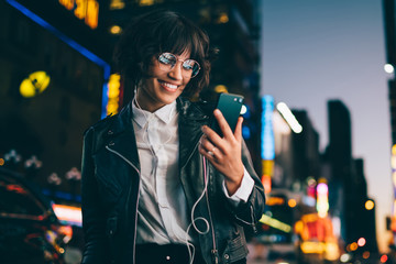 Happy influence blogger in classic glasses and trendy leather jacket reading funny social publication during mobility networking on cellphone, millennial hipster girl listening positive music