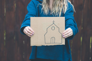 Person in a blue long sleeve shirt holding a cardboard sign with a church drawn on it.