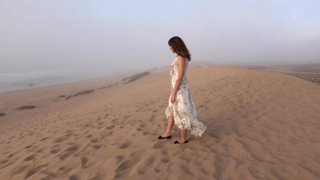 Young woman walking along sea shore sand beach in fog rainy day. Girl in long dress enjoy view on sand dune near ocean in Morocco