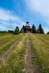 Old Russian wooden church, chapel on a field. With blue sunny sky, road and forest on background.