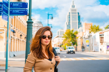 A young woman in sunglasses stands at a traffic light on a business street in Moscow on a summer day