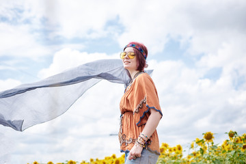 Young woman with red burgundy hair, wearing boho hippie clothes, holding grey transparent shawl scarf, waving on wind on yellow sunflowers field. Creative female portrait on natural background.