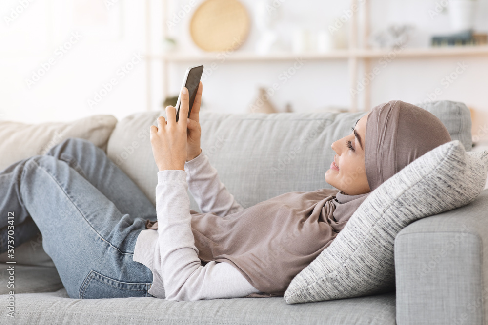 Wall mural happy arabic woman in hijab lying on couch at home using smartphone