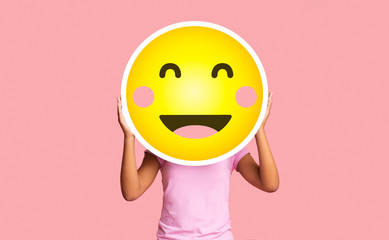 Black girl hiding face behind happy shy emoji emoticon on pink background