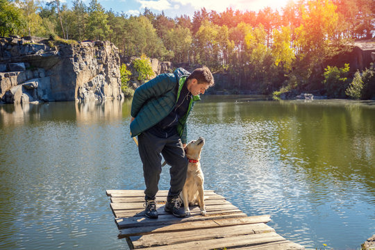 A Man With Labrador Retriever Dog Stands On A Wooden Deck On The Beautiful Granite Lakeshore