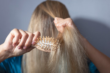 blonde combs dry hair with a wooden comb. On a gray background. Hair disease, fungus, brittle hair, dry scalp. scalp hair damage