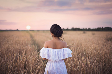 Portrait of a young beautiful brunette girl in a white summer dress standing in a field with wheat ears on the background of the sunset sky