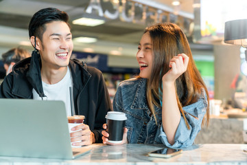 Happy asian couple take off face mask in a coffee shop surfing internet on laptop. Young man and woman in a restaurant looking at touch screen computer laugh smile together