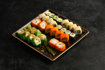 Large maki sushi set with a varied assortment of Japanese rolls in a square plate on a black stone table.