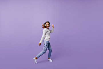 Fototapeta na wymiar Dancing Slavic female student in stylish jeans and winter sweater moves on purple background. Brunette with headphones poses for full-length photo