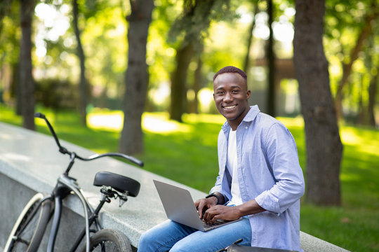 Joyful African American guy with bike working on laptop online at urban park, free space