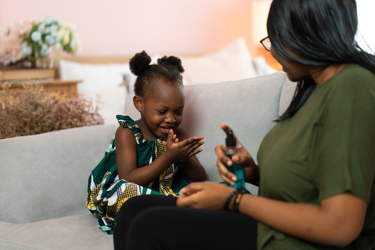 Young African American mother use spray sanitizer on hands child in a protective mask in living room at home. Black lives matter concept. Protect against coronavirus - Powered by Adobe