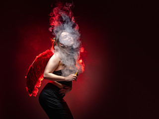 Girl with a VAPE on a red background. A woman in a cloud of smoke from an electronic cigarette. The girl poses and smokes a VAPE. A Vaper with red wings. Vaping fantasy.