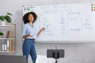 Fotobehang Online lesson for students. Smiling girl points at blackboard and teaches english at webcam © Prostock-studio