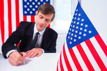 An American official issues a permit. A man writes against the background of USA flags. Public service in America. Registration of documents for a trip to the United States of America. US immigration.