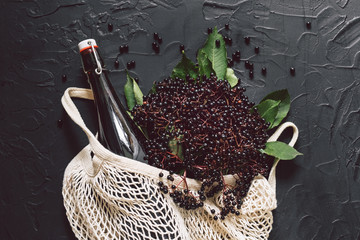 A bottle of elderberry syrup on a dark background with fresh elderberries and string bag